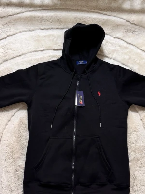 Svart hoodie från Polo Ralph Lauren - Svart hoodie med dragkedja från Polo Ralph Lauren. Klassisk design med huva och snören, två fickor framtill och ikonisk röd logobrodyr på bröstet. Perfekt för en avslappnad och stilren look. Passar mer som M
