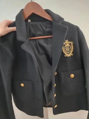 Svart kavaj med guldknappar och emblem - Stilren svart kavaj med två stora fickor framtill, dekorerad med guldknappar och ett broderat emblem på bröstet. Kavajen har klassisk krage och är helfodrad för extra komfort. Perfekt för en preppy look och passar till många olika outfits.