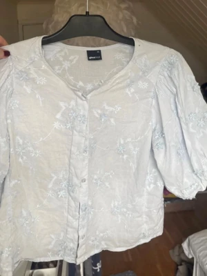 Ljusblå broderad blus från Gina Tricot - Supersöt ljusblå blus från Gina Tricot med puffiga korta ärmar och fina blommiga broderier. Blusen har knappar framtill och en avslappnad passform. Perfekt till jeans eller kjol för en fräsch look. Nypris ca 400kr. Endast använd ca 2-3 gånger.🩵