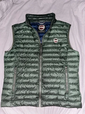 Mörkgrön dunväst från Colmar - Stilren mörkgrön dunväst från Colmar med quiltad design och hög krage. Västen har två dragkedjeförsedda fickor framtill och en inneficka, Colmar-logga på bröstet samt på baksidan. Inge Defekter