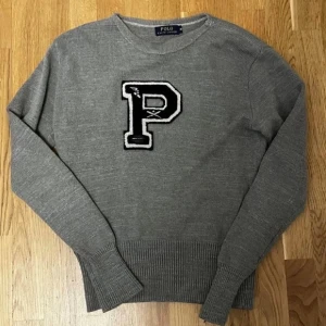 Stickad Ralph Lauren Tröja - Grå stickad tröja från Polo Ralph Lauren med svartvit P-applikation. Klassisk college-stil med rund hals och ribbade muddar. Midjan har krympt något – perfekt för slim fit och passar även Small.