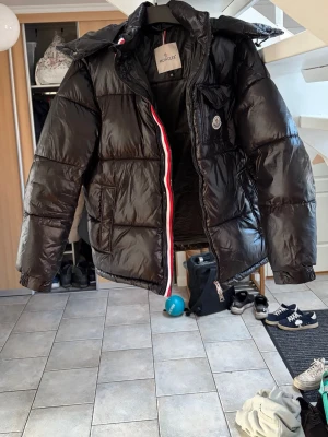 Svart dunjacka från Moncler - Snygg svart dunjacka från Moncler med glansig finish och klassisk Moncler-logga på bröstet. Jackan har dragkedja med röd, vit och blå detalj, två sidofickor och en huva. Perfekt för kalla vinterdagar och riktigt trendig stil. Köpt i vinter, dragkedjan på ena fickan är sönder 