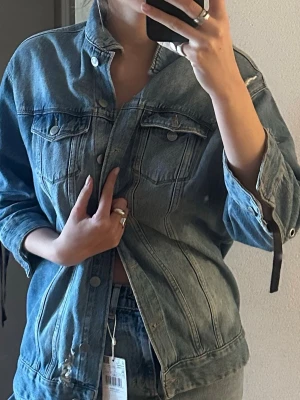  blå jeansjacka med svart snörning  - Snygg blå jeansjacka med raka sömmar och två bröstfickor med lock och knapp. Jackan har en avslappnad passform och knäpps framtill med metallknappar. Perfekt att slänga över en t-shirt för en cool och tidlös look. Från gina tricot