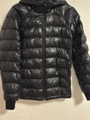 Svart dunjacka Peak Performance - PRIS KAN DISKUTERAS. Snygg svart dunjacka från Peak Performance med quiltad design och huva. Jackan har dragkedja framtill, två sidofickor och ribbade muddar vid ärmsluten. Märkeslogga på ärmen ger en sportig touch. Perfekt för kalla vinterdagar.