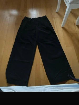 Svarta vida kostymbyxor - Snygga svarta kostymbyxor med vida ben. Byxorna har pressveck framtill, sidofickor och bälteshällor i midjan. Stängs med knapp och dragkedja. Perfekta för en clean och stilren look.