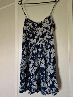 Blommig marinblå kortklänning - Dekorativ snörningen i ryggen och reglerbara axelband. Klänningen är använd ett fåtal tillfällen och har inga skavanker vad jag har kunnat se. Stl M 
