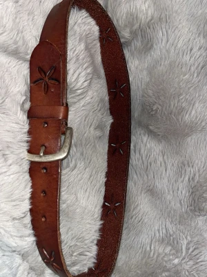 Brunt skärp med blommönster - Säljer ett brunt skärp i skinn med blommönster som är stansade längs hela bandet. Skärpet har ett klassiskt spänne i silverfärg och en rustik look som passar till jeans eller shorts. Perfekt accessoar för att lyfta din outfit.