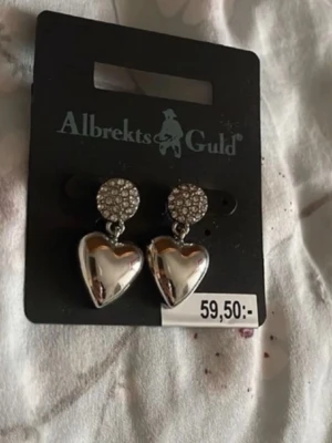 Silverfärgade hjärtörhängen Albrekts Guld - Snygga örhängen i silverfärg från Albrekts Guld med glittrande runda toppar täckta av små stenar och hängande blanka hjärtan. Perfekta för att ge din look lite extra bling och romantisk vibe.