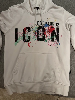 Vit hoodie Dsquared2 ICON - Säljer en vit hoodie från Dsquared2 med stor svart 'ICON'-text och färgglada färgstänk på bröstet. Hoodien har huva med dragsko och en stor magficka. Perfekt för dig som vill sticka ut med en streetstyle-look. Sparsamt använd, Skick 9/10