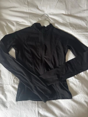 Svart ziptröja från Gina Tricot - Snygg svart ziptröja från Gina Tricot med figurnära passform och hög krage. Tröjan har långa ärmar och dragkedja framtill, perfekt för en sportig och stilren look. 