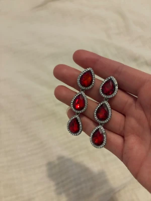 Röda droppformade örhängen med kristaller - Säljer ett par eleganta örhängen med stora röda droppformade stenar omgivna av gnistrande små kristaller. Örhängena har en silverfärgad infattning och är riktigt statement med flera hängande delar. Perfekt för dig som vill sticka ut! Örhängen är ifrån glitter och aldrig tyvärr kommit till användning