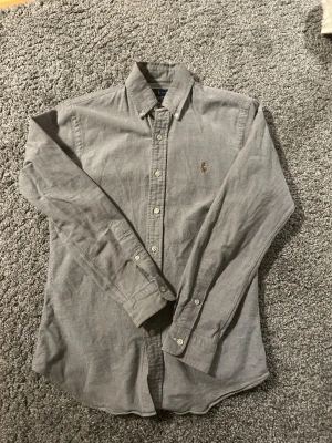 Grå skjorta från Ralph Lauren - Snygg grå skjorta från Ralph Lauren i modellen Slim Fit Stretch Oxford. Jag skulle säga att skjortan skiftar i blått och grått. Skjortan har button-down krage, långa ärmar och klassisk broderad logga på bröstet. Skjortan är i bra skick och är riktigt snygg.