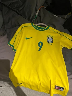 Brasilien Ronaldo 9 matchtröja Nike - Gul Brasilien fotbollströja från Nike med gröna och blå detaljer, nummer 9 och Ronaldo på ryggen. Klassisk landslagströja med broderad Nike-logga och Brasilien-emblem på bröstet. Perfekt för dig som älskar fotboll och vill sticka ut på planen.