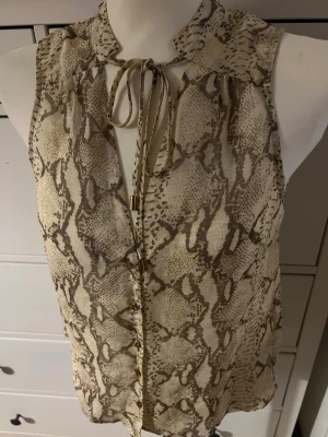 Beige ärmlös blus med ormmönster - Säljer en snygg ärmlös blus i beige med ormmönster och knytband vid halsen. Blusen har en luftig passform och tunnt material som känns skönt mot huden. Perfekt för dig som vill ha en trendig och unik topp till din garderob. Tyvärr är storlekslappen bortklippt då jag köpte den så