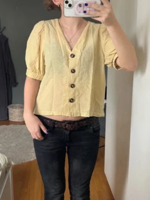 Gul blus med knappar och puffärm - Snygg ljusgul blus med v-ringning och stora bruna knappar framtill. Blusen har puffiga korta ärmar med resår och är tillverkad i ett lätt, strukturerat tyg som ger en avslappnad look. Perfekt att matcha med jeans för en cool stil.