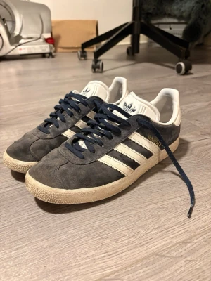 Adidas Gazelle mörkblå sneakers - Säljer ett par klassiska Adidas Gazelle sneakers i mörkblå mocka med vita detaljer och vita ränder på sidorna. Skorna har mörkblå snören, vit plös med Adidas-logga och vit sula. Perfekta för dig som gillar retrostil och streetwear.