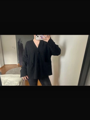 Svart oversized kofta med fickor - Säljer en svart oversized kofta med v-ringning och stora framfickor. Koftan har knäppning framtill och långa ärmar med avslappnad passform. Perfekt för lager på lager och enkel att styla till olika outfits.