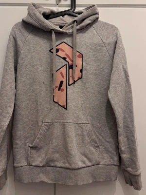 Grå hoodie med grafiskt tryck (peak peformance) - Snygg grå hoodie med huva och dragsnören. På bröstet finns ett stort grafiskt tryck i rosa och svart med mönster. Hoodien har en klassisk känguruficka framtill och ribbade muddar vid ärmslut och nederkant. Perfekt för en avslappnad streetstyle-look.