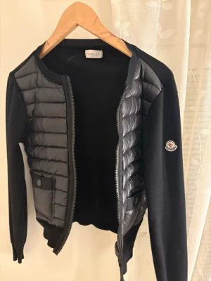 Svart cardiganjacka från Moncler strl S - Snygg svart cardiganjacka från Moncler med quiltade partier framtill och på ärmarna. Jackan har dragkedja, rund halsringning och en Moncler-logga på vänster ärm. Materialmix med stickade partier och vadderade detaljer ger en modern look.