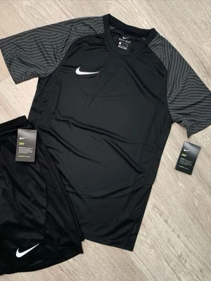 Svart träningsset från Nike - Snyggt svart träningsset från Nike med t-shirt och shorts. T-shirten har gråmönstrade ärmar och vit Nike-logga på bröstet. Shortsen är helsvarta med vit logga. Tillverkade i lätt och snabbtorkande Dri-FIT material som andas.