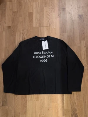 Svart långärmad topp Acne Studios - Svart långärmad topp från Acne Studios med vit text 'Acne Studios STOCKHOLM 1996' på bröstet. Klassisk rund halsringning och relaxed fit. Perfekt till jeans eller kjol för en clean och modern look.