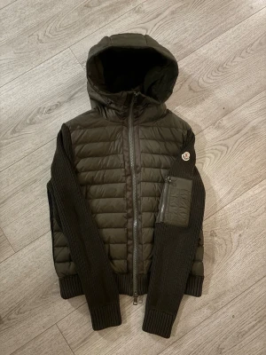 Mörkgrön Moncler pufferjacka med huva - Säljer en mörkgrön pufferjacka från Moncler med stickade ärmar och ribbade muddar. Jackan har huva, dragkedja framtill och en ficka med dragkedja på ena ärmen. Klassisk Moncler-logga på ärmen. Perfekt för kalla vinterdagar.