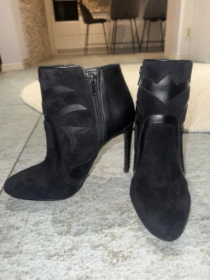 Svarta stövletter med klack - Snygga svarta stövletter med spetsig tå och hög smal klack. Skorna har en mix av mocka och glansigt skinn, samt dekorativa detaljer i lädret på sidorna. Dragkedja på insidan för enkel påtagning. Perfekta för dig som vill ha en edgy look. Aldrig använda 