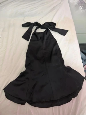 Svart halterneckklänning Gina Tricot - Supersnygg svart halterneckklänning från Gina Tricot Young. Klänningen har en bred knytning i nacken och är tillverkad i ett glansigt polyestertyg som ger en elegant look. Perfekt för dig som vill ha en stilren och trendig outfit.