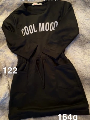 Svart klänning med tryck och snörning - Svart klänning med texten 'COOL MOOD' i vitt framtill. Klänningen har lång ärm, snörning i midjan och två fickor framtill. Perfekt för en avslappnad och cool stil.🌸 Oanvänd. Storlek 122