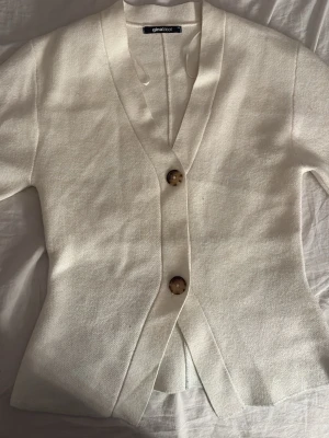 Vit kofta från Gina Tricot - Stilren vit kofta från Gina Tricot med två stora, bruna knappar framtill. Koftan har v-ringning, korta ärmar och är gjord i ett mjukt, stickat material. Perfekt att slänga över en topp för en enkel men snygg look. Lite färg från jeansen på sista bilden men går bort i tvätten.
