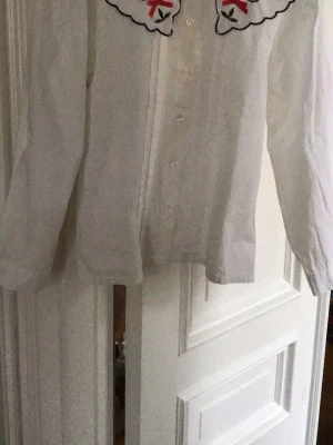 Vit skjorta med broderad krage - Vit skjorta med lång ärm och knappar framtill. Kragen har broderade blommor och röda rosetter, vilket ger en unik och söt detalj. Skjortan är i bomull och har en klassisk passform. Perfekt för dig som gillar romantiska detaljer.