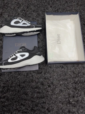 Dior B30 svarta och vita sneakers - Snygga Dior B30 sneakers i svart och vitt med stora reflekterande detaljer på sidorna och meshpaneler för extra andningsförmåga. Tjock vit sula och vita skosnören ger en modern vibe. Perfekta för dig som vill sticka ut med exklusiva och trendiga skor.