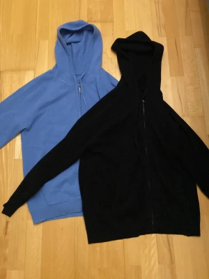 Kashmir hoodies - Två hoodies i olika färger, en blå och en svart, båda med dragkedja framtill och huva. Hoodiesarna har en klassisk passform och ribbade muddar vid ärmar och nederkant. Perfekta för en avslappnad och stilren look.