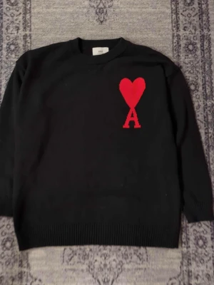 Svart stickad tröja från Ami Paris - Snygg svart stickad tröja från Ami Paris med deras ikoniska röda hjärta och bokstaven A broderat på bröstet. Tröjan har rund halsringning, långa ärmar och ribbade muddar. Perfekt för dig som gillar stilrena plagg med en twist.