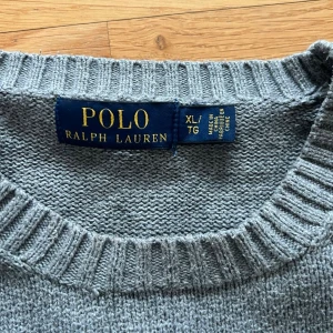 Grå stickad tröja från Polo Ralph Lauren - Klassisk grå stickad tröja från Polo Ralph Lauren med rund halsringning och ribbade muddar vid ärmslut och nederkant. Tröjan har en diskret broderad logga på bröstet och är tillverkad i mjukt material som passar perfekt till lager på lager.