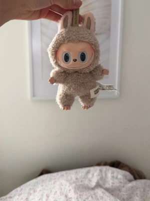 Äkta Macaron Labubu - Söt beige nyckelring från Popmart i mjuk plysch med gulliga öron och storögd ansikte. Perfekt att hänga på väskan eller nyckelknippan för en personlig touch. Tillverkad i mjukt material och har en praktisk metallring. Köpt på Amart!!