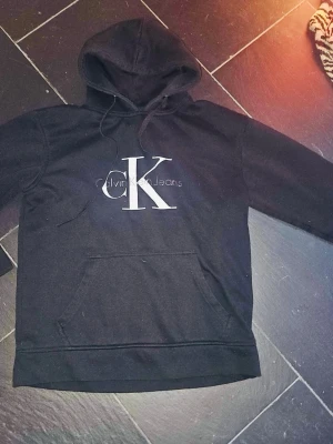 Svart hoodie från Calvin Klein Jeans - Säljer en svart hoodie från Calvin Klein Jeans med stor CK-logga på bröstet. Hoodien har huva med dragsko och en klassisk magficka. Materialet är mjukt och bekvämt, perfekt för en avslappnad look.