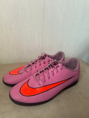 Rosa Nike Mercurial fotbollsskor - Säljer ett par rosa Nike Mercurial fotbollsskor med svart och orange swoosh. Skorna har låg profil, snörning och slitstark gummisula för spel på konstgräs eller inomhus. Snygg design som sticker ut på planen och passar perfekt för dig som vill ha stil och prestanda.