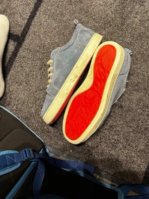 Ljusblå Louboutin sneakers med röd sula - Snygga sneakers från Christian Louboutin i ljusblå mocka med beigea skosnören och ikonisk röd sula. Låg modell med rund tå och diskreta sömmar. Perfekta för dig som vill sticka ut med exklusiva detaljer och klassisk design.