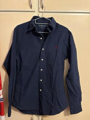 Mörkblå skjorta från Ralph Lauren - Klassisk mörkblå skjorta från Ralph Lauren med knappar framtill och röd broderad logga på bröstet. Skjortan har lång ärm, normal passform och är tillverkad i mjuk bomull. Perfekt för en stilren och avslappnad look. Kom med prisförslag.