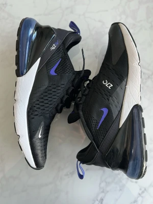 Nike Air Max 270 svarta sneakers - Snygga Nike Air Max 270 sneakers i svart mesh med vita och blå detaljer. Skorna har en tydlig blå swoosh, stor synlig Air-enhet i hälen och ventilerande ovandel. Perfekta för dig som gillar sportig och modern stil.