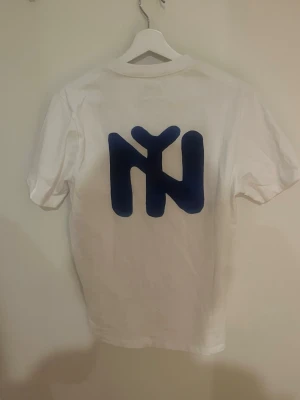 Vit Aimé Leon Dore t-shirt - Säljer en vit t-shirt från Aimé Leon Dore med tryck i svart på bröstet och en stor blå grafisk logga på ryggen. Klassisk passform och rund hals. Perfekt för dig som gillar streetwear och stilrena plagg.