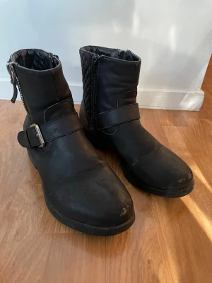 Svarta fodrade vinterskor med spänne - Snygga svarta fodrade stövlar i konstläder med dekorativt spänne och dragkedja på sidan. Bootsens insida är quiltad för extra komfort och de har en robust sula som passar till många olika stilar.De har sitt slitage på ytan men bra invändigt. 