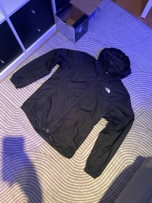 Svart vindjacka från The North Face - Snygg svart vindjacka från The North Face med huva och dragkedja framtill. Jackan har en minimalistisk design med logga på bröstet och är tillverkad i lätt, vindtätt material. Perfekt för blåsiga dagar och enkel att packa ner i väskan.