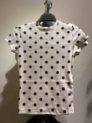 prickig topp - Vit topp med svarta prickar i ett lekfullt mönster. Modellen har rund hals, korta ärmar och en figurnära passform. Tillverkad i mjuk bomull som känns skön mot huden. Perfekt för dig som vill ha en trendig och enkel look.