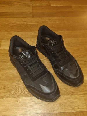 Valentino rockrunners - Säljer ett par helsvarta sneakers från Valentino Garavani med klassisk låg siluett. Skorna har en mix av mocka och läderdetaljer, svarta skosnören och diskret logga på plösen. Perfekta för dig som gillar stilrena och exklusiva sneakers. Storlek 46 sitter mer som 44~ bra skick inga defekter 