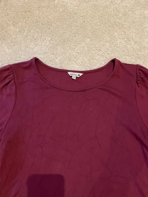 Vinröd topp från Indiska - Enkel och snygg vinröd topp från Indiska med rund halsringning och lätt puffade axlar. Tillverkad i mjukt material som känns skönt mot huden. Perfekt att styla med jeans eller kjol för en trendig look.