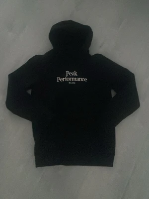Svart hoodie från Peak Performance - Säljer en svart hoodie från Peak Performance med vit logga och text på bröstet. Klassisk modell med huva och ribbade muddar. Perfekt för dig som gillar stilrena och bekväma plagg. Materialet känns mjukt och skönt mot huden. Storlek: 160 13/14 år, storleken fanns inte men den motsvarar XS