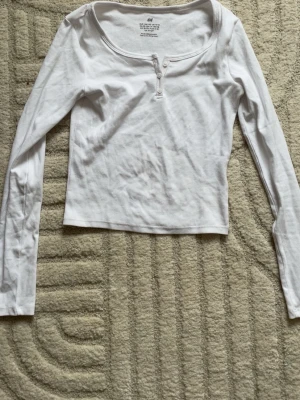 Vit långärmad topp från H&M - Säljer en vit långärmad topp från H&M med rundad urringning och knappdetalj framtill. Toppen är croppad och har en slim passform som sitter snyggt på kroppen. Perfekt att styla med jeans eller kjol för en clean look.