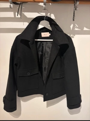 Svart kort jacka från Nelly - Stilren svart kort jacka från Nelly med stora fickor framtill och knappar som detalj. Jackan har bred krage, lång ärm och är fodrad på insidan. Perfekt för dig som gillar en clean och modern look.Tunnare jacka från Nelly, aldrig använd med prislapp kvar, stl xxs/32